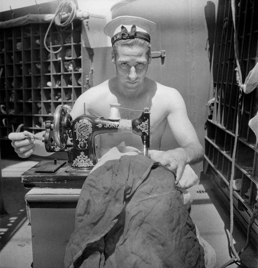 Cecil Beaton (English, 1904-1980) 'A Royal Navy sailor on board HMS Alcantara uses a portable sewing machine to repair a signal flag during a voyage to Sierra Leone' March 1942