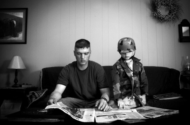 Andrea Bruce (American, b. 1973) 'Untitled [A father home on leave reading the newspaper with his son, who wears his dad’s helmet]' 2006