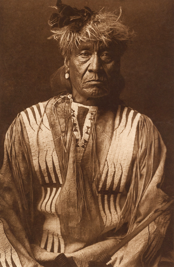 Edward S. Curtis (American, 1868-1952) '[Atsina Indian, Red Whip, half-length portrait, seated, facing front, wearing feather, beaded buckskin shirt, holding pipe in left hand]' c. 1908