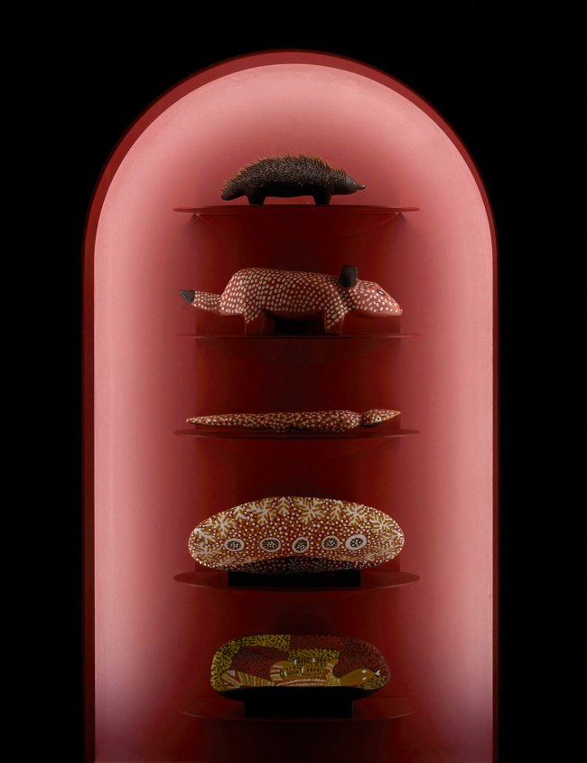 Installation view of the exhibition '1,001 Remarkable Objects' at Powerhouse Ultimo, Sydney showing at top, Carved form, Echidna shape, bean wood / echidna quills, painted, Louie Pwerle, Ngkawenyerre camp, Utopia, Northern Territory, Australia, 1996; and at second top, Figure, dog, Devil Dog, wood, Billy Petyarre, Utopia Station, Northern Territory, Australia, 1991; at second bottom, Coolamon, Bush Tomato Dreaming, wood / acrylic paint, Billy Petyarre, Utopia Station, Northern Territory, 1990-1991