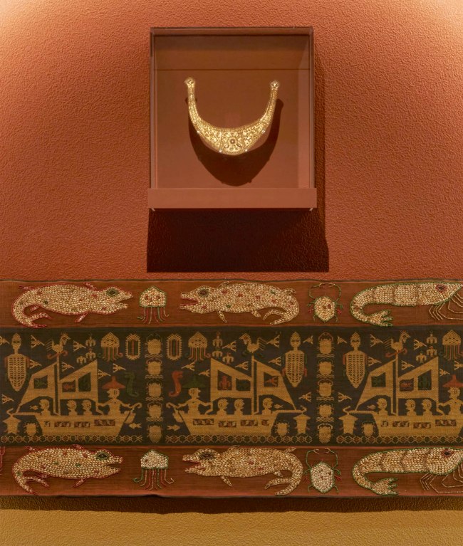 Installation view detail of the exhibition '1,001 Remarkable Objects' at Powerhouse Ultimo, Sydney showing at top, Man's ceremonial headpiece (lamba), sheet gold alloy, Sumba, Indonesia, 1880-1950; and at second top, Textile length (mens waist cloth), handwoven cotton / shells / beads, maker unknown, East Sumba, Indonesia, 2000-2004