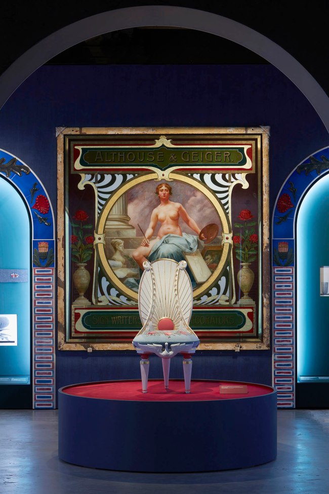 Installation view of the exhibition '1,001 Remarkable Objects' at Powerhouse Ultimo, Sydney showing in the background, Painting, 'Althouse & Geiger / Sign Writers, Painters, Decorators', glass / oil / gold leaf, designed and made by Althouse and Geiger Pty Ltd, Sydney, New South Wales, Australia, 1895-1905; and in the foreground Chair, reproduction, based on Lucien Henry, 'Lyrebird' or 'staghorn fern' design, jelutong & kauri timber / gesso finish / needlework, made by International Conservation Services Pty Ltd, Australia, 2000