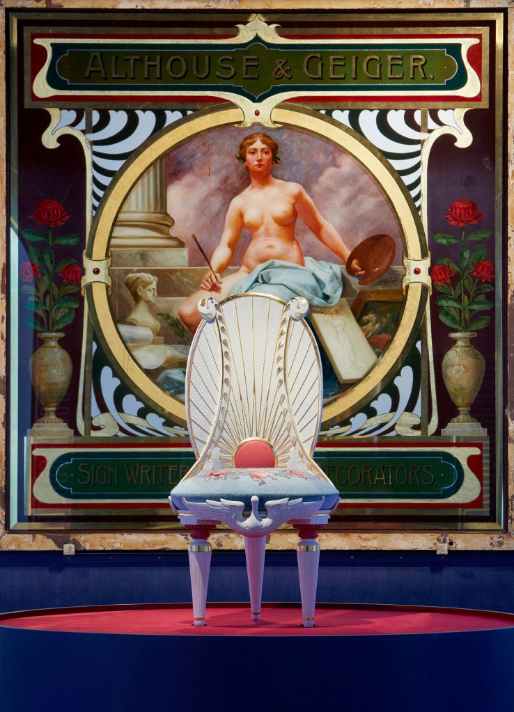 Installation view detail of the exhibition '1,001 Remarkable Objects' at Powerhouse Ultimo, Sydney showing in the background, Painting, 'Althouse & Geiger / Sign Writers, Painters, Decorators', glass / oil / gold leaf, designed and made by Althouse and Geiger Pty Ltd, Sydney, New South Wales, Australia, 1895-1905; and in the foreground Chair, reproduction, based on Lucien Henry, 'Lyrebird' or 'staghorn fern' design, jelutong & kauri timber / gesso finish / needlework, made by International Conservation Services Pty Ltd, Australia, 2000